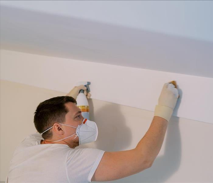 A man wearing protective gear, cleaning mold off a white wall.