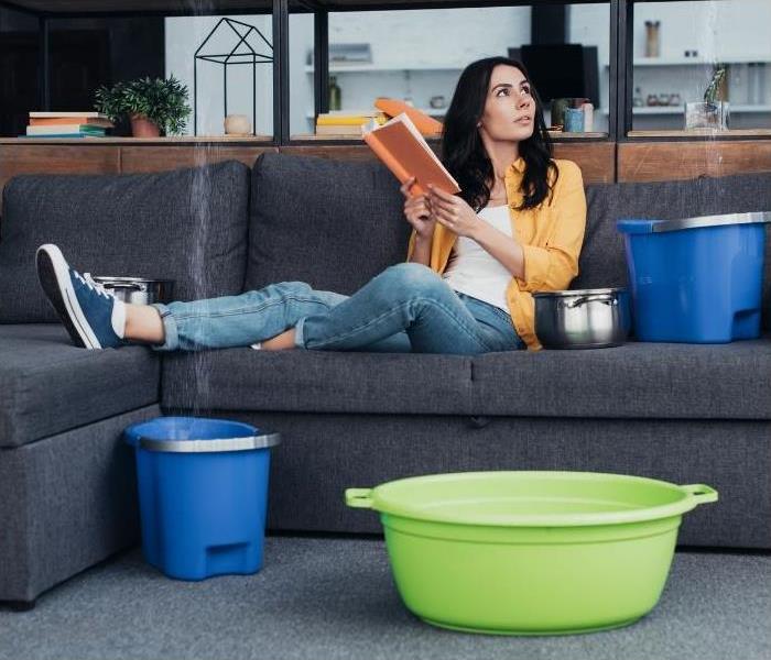 Woman reading a book while laying on a couch. There are three buckets surrounding her, catching water as it leaks.