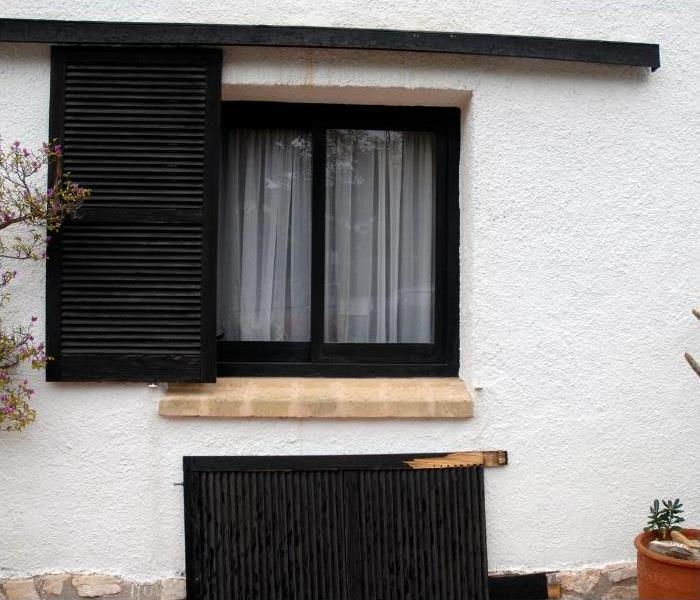An exterior window with one storm shutter installed and a second one laying on the ground, propped up on the house.